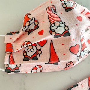 May include: Pink fabric face mask with a repeating pattern of red gnomes, hearts, and candy canes.