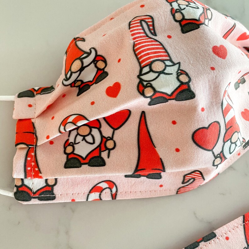 May include: Pink fabric face mask with a repeating pattern of red gnomes, hearts, and candy canes.