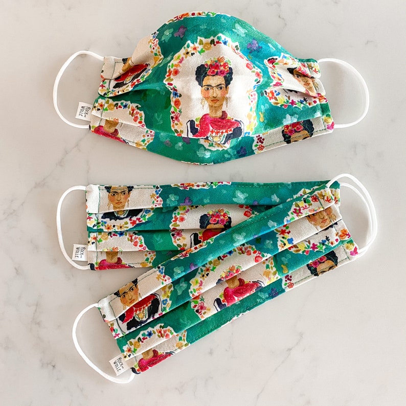 May include: Two green and white face masks with a floral pattern and a portrait of Frida Kahlo. The masks are pleated and have white elastic ear loops. The text "Rock the Vote" is printed on the masks.