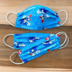 Op de afbeelding: Twee blauwe stoffen mondmaskers met witte elastische oorlussen. De maskers hebben een herhalend patroon van Sonic the Hedgehog-personages en sterren. De tekst "Sonic" staat op beide maskers.