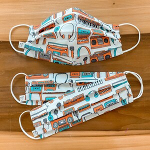 May include: Two pleated face masks featuring a white background with a repeating pattern of orange and teal retro music equipment, including boomboxes, cassette tapes, microphones, and headphones. Each mask has white elastic ear loops and a small tag.