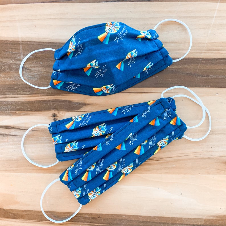 May include: Two blue fabric face masks with white elastic ear loops. The masks are printed with a repeating pattern of the Millennium Falcon spaceship and the words "Millennium Falcon".