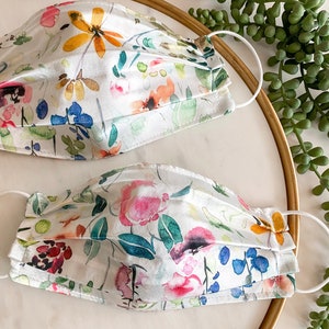 May include: Two white face masks with a floral print. The masks are made of a soft, silky fabric and have a pleated design. The floral print features watercolor-style flowers in shades of pink, blue, yellow, and green.