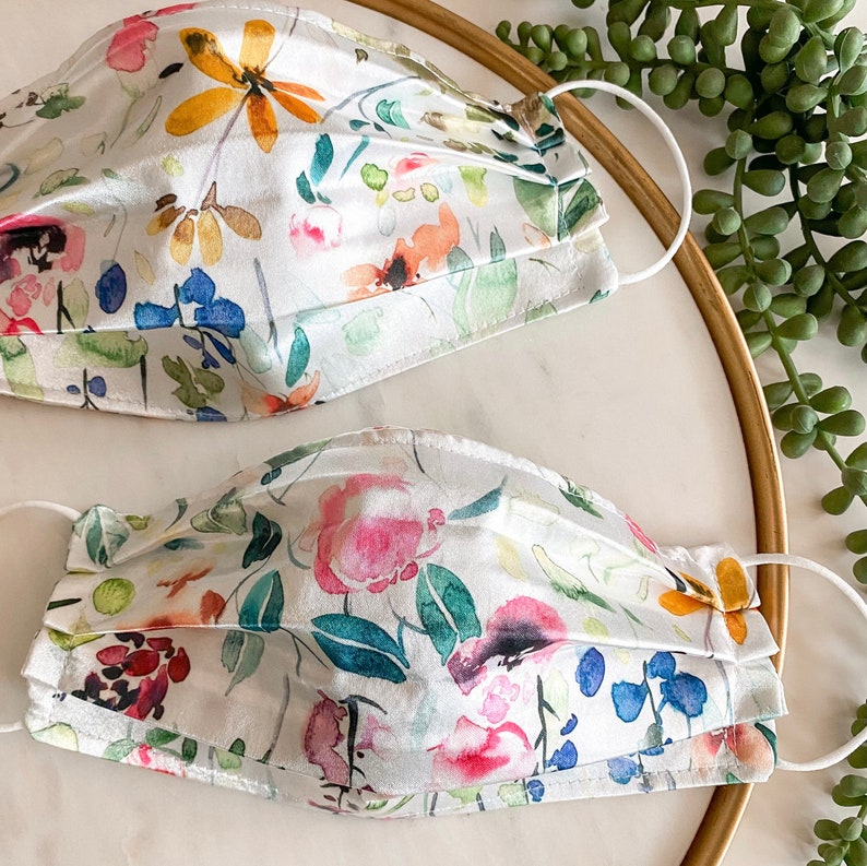 May include: Two white face masks with a floral print. The masks are made of a soft, silky fabric and have a pleated design. The floral print features watercolor-style flowers in shades of pink, blue, yellow, and green.
