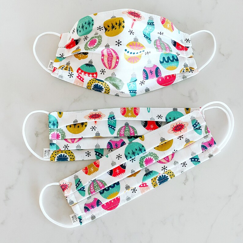 May include: Two white face masks with a colorful Christmas ornament pattern. The masks are pleated and have adjustable ear loops. The text "WOLLE" is printed on the bottom of the masks.