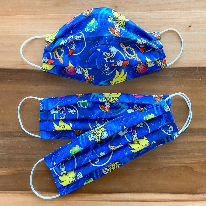 Op de afbeelding: Drie blauwe stoffen mondmaskers met witte elastieken. De maskers hebben een herhalend patroon van Sonic the Hedgehog-personages in verschillende poses.