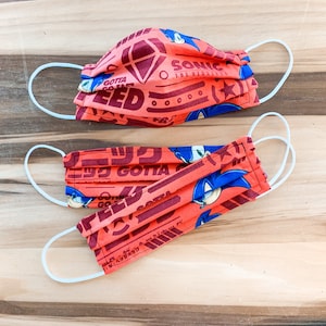 Op de afbeelding: Twee rode en blauwe stoffen mondmaskers met een Sonic the Hedgehog-thema. De maskers hebben de tekst "Gotta Go Fast" en "Sonic The Hedgehog" erop gedrukt.