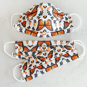May include: Three white face masks with a repeating pattern of orange butterflies and floral designs. The masks are made of fabric and have adjustable ear loops.