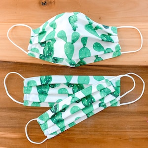May include: Two face masks with a white background and green cactus print. The larger mask has a single layer of fabric, while the smaller mask has three layers of fabric.