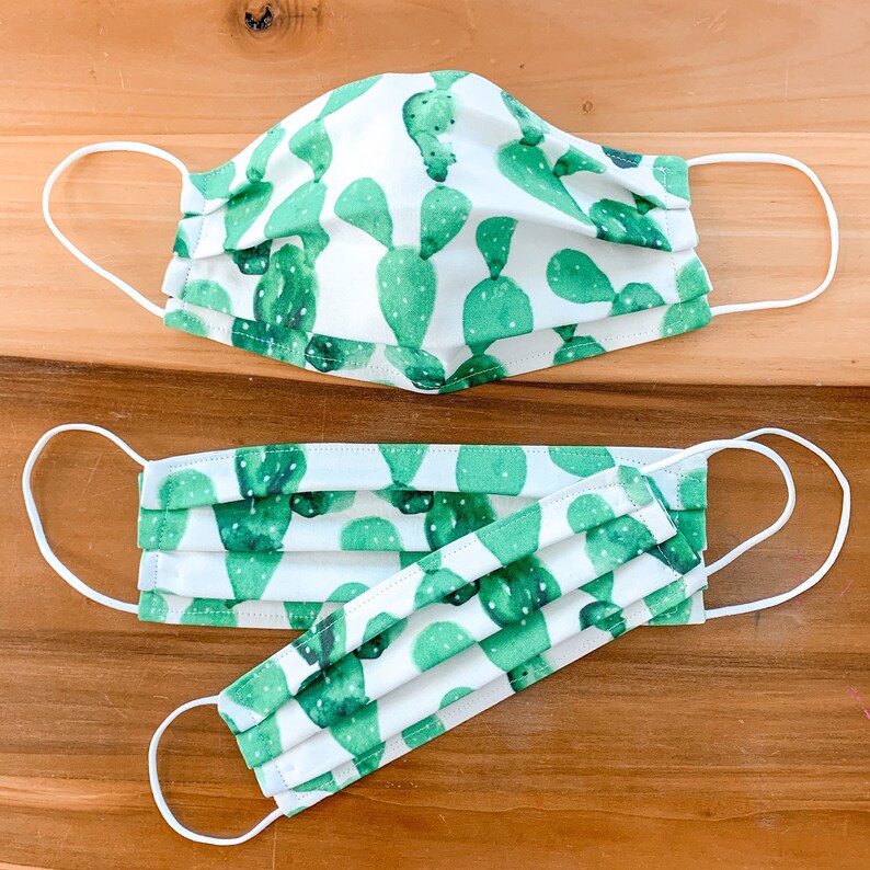 May include: Two face masks with a white background and green cactus print. The larger mask has a single layer of fabric, while the smaller mask has three layers of fabric.