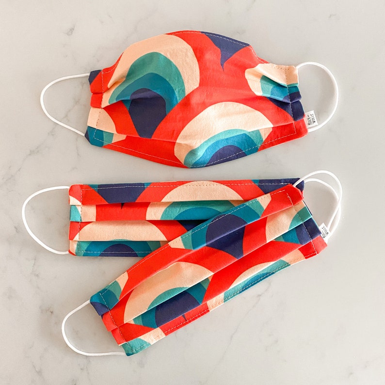 May include: Two face masks with a colorful geometric pattern in shades of red, blue, and green. The masks are made of fabric and have white elastic ear loops.