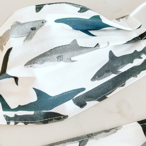 May include: White fabric face mask with a repeating pattern of gray and blue sharks. The sharks are in various sizes and positions.