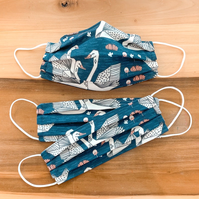 May include: Two teal blue fabric face masks with white swan designs and white elastic ear loops. The masks are pleated and have a floral pattern.