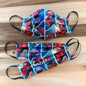 May include: Two face masks with a Spiderman theme. The masks are made of fabric and have a pleated design. The top mask has a red, blue, and black color scheme. The bottom mask has a red, black, and white color scheme.