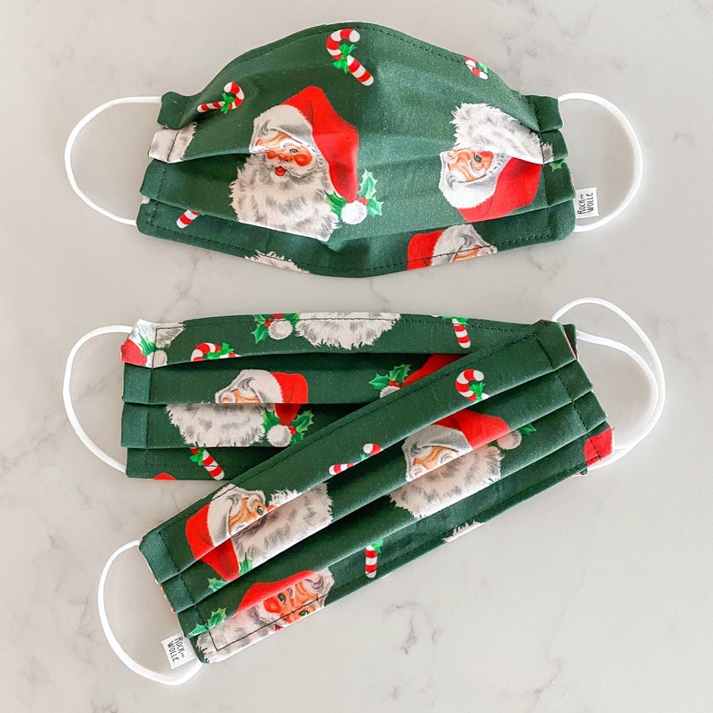 May include: Two green fabric face masks with a repeating pattern of Santa Claus and candy canes. The masks are pleated and have white elastic ear loops.