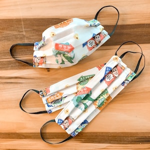 May include: Two white fabric face masks with a Toy Story theme. The masks feature characters like Buzz Lightyear, Woody, and Rex. The masks have a white background with colorful cartoon characters and text that says "Hey, Howdy, Hey!" and "Buzz".