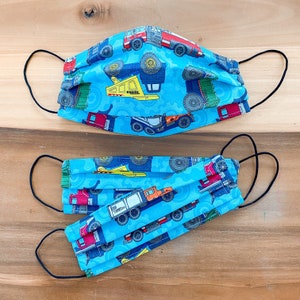 May include: Two blue fabric face masks with a repeating pattern of construction vehicles. The masks are pleated and have adjustable ear loops. The top mask is folded in half, while the bottom mask is fully extended.