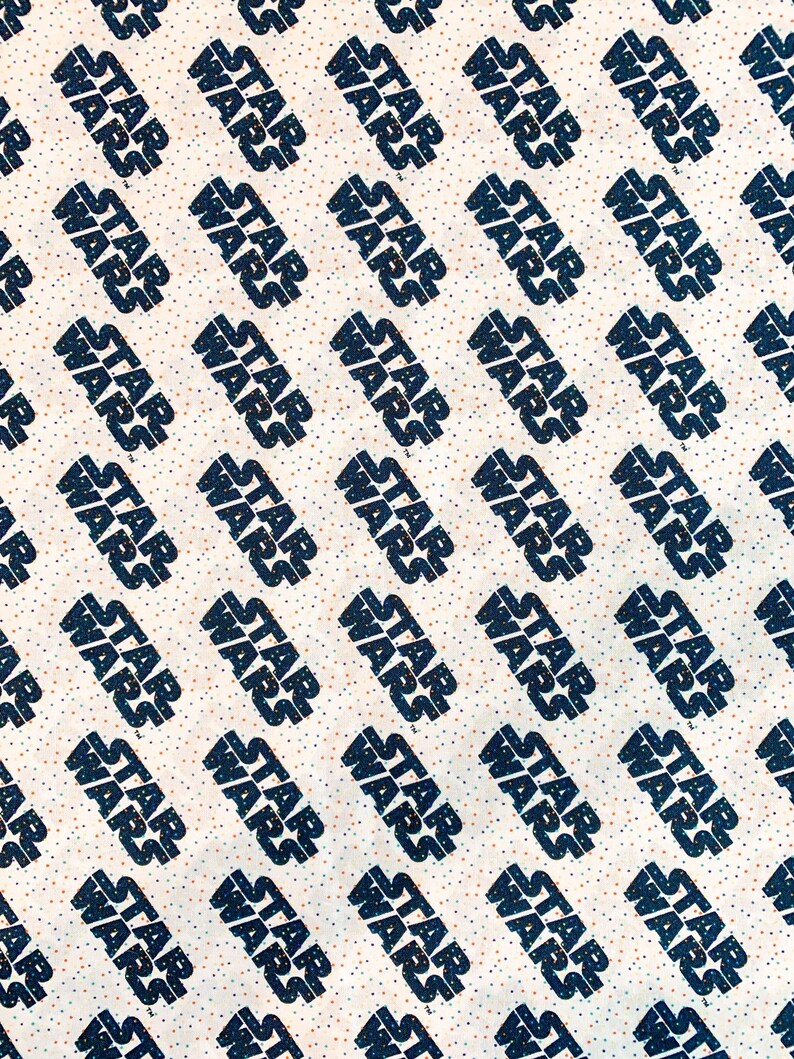 May include: White fabric with a repeating pattern of the words "Star Wars" in black text. The background is a light beige with small black and orange dots.