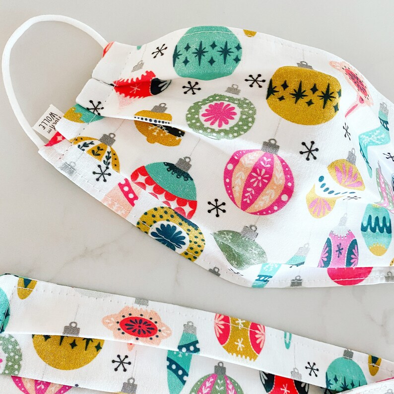 May include: A white fabric face mask with a repeating pattern of colorful Christmas ornaments. The ornaments are in shades of red, green, yellow, pink, and blue. The mask has a white elastic band.