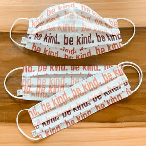 May include: Two white face masks with elastic ear loops. The masks are made of a soft, white fabric with a repeating pattern of the words "be kind" in shades of pink and brown.