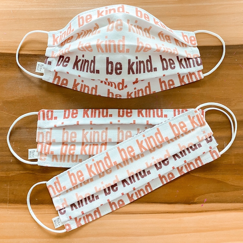 May include: Two white face masks with elastic ear loops. The masks are made of a soft, white fabric with a repeating pattern of the words "be kind" in shades of pink and brown.