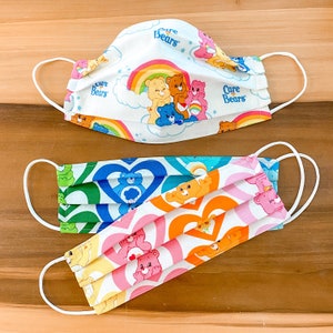 May include: Three colorful face masks with white elastic ear loops. The top mask is white with a Care Bears rainbow design. The other two masks have heart and Care Bear patterns in green, blue, orange, and pink. The masks are pleated.