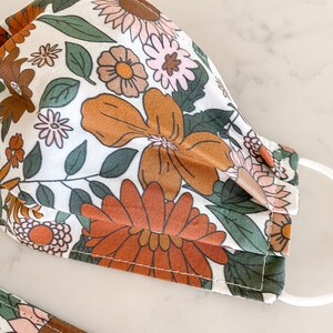 Peut inclure: Un masque facial en tissu blanc avec un motif floral dans les tons orange, marron, rose et vert. Le masque a un design pliss&eacute; et des boucles d'oreille &eacute;lastiques blanches.