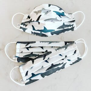 May include: Two white fabric face masks with a repeating pattern of gray and blue sharks. The masks have white elastic ear loops.