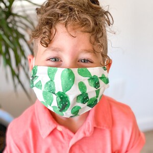 May include: A young person wearing a white face mask with a green cactus print. The mask has pleats and is covering the person's nose and mouth. The person is wearing a pink polo shirt.