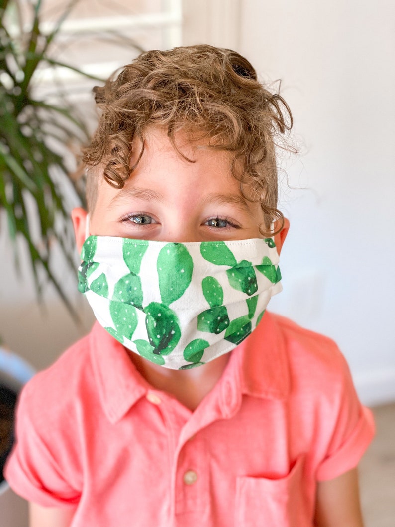 May include: A young person wearing a white face mask with a green cactus print. The mask has pleats and is covering the person's nose and mouth. The person is wearing a pink polo shirt.