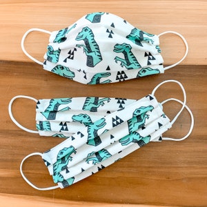 May include: Two white face masks with a repeating pattern of green dinosaurs and black triangles. The masks have white elastic ear loops.