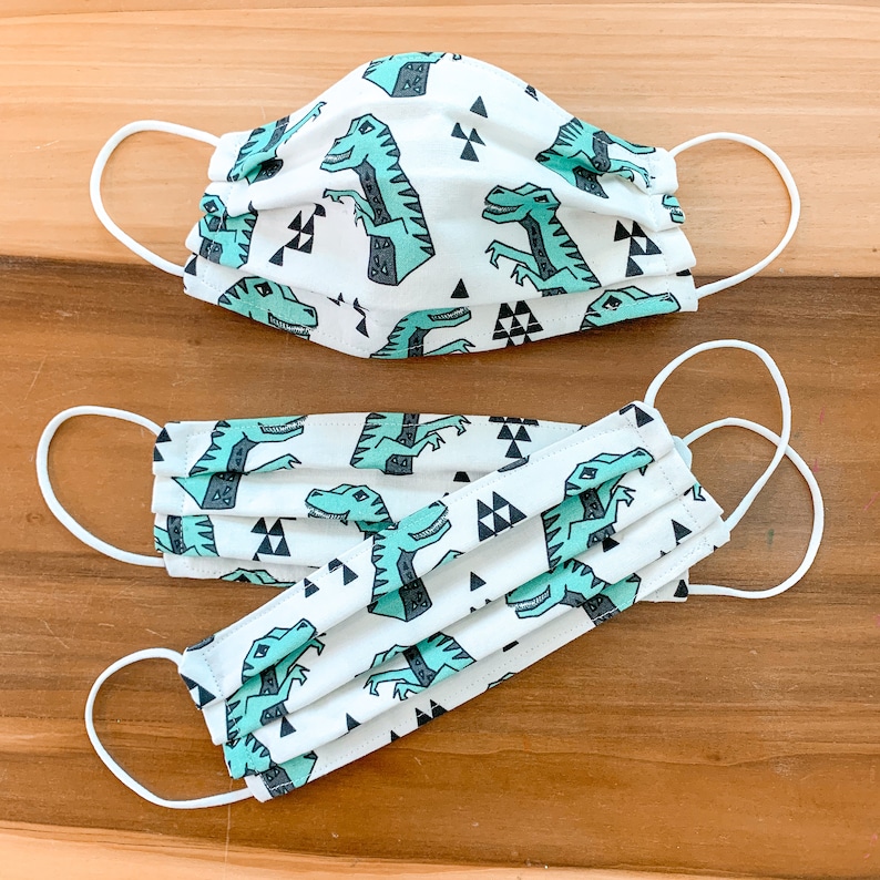 May include: Two white face masks with a repeating pattern of green dinosaurs and black triangles. The masks have white elastic ear loops.