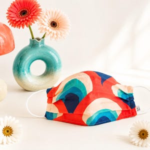 May include: A fabric face mask featuring a repeating pattern of red, navy blue, teal, and cream-colored arches. The mask has white elastic ear loops and a small white tag on the side. It is positioned near a teal vase and a red mushroom-shaped object.