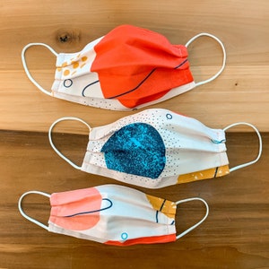 May include: Three fabric face masks with abstract patterns. The masks are white with different colored geometric shapes and lines. The masks are made of cotton and have elastic ear loops.