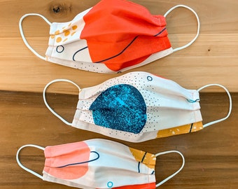 May include: Three fabric face masks with abstract patterns. The masks are white with different colored geometric shapes and lines. The masks are made of cotton and have elastic ear loops.