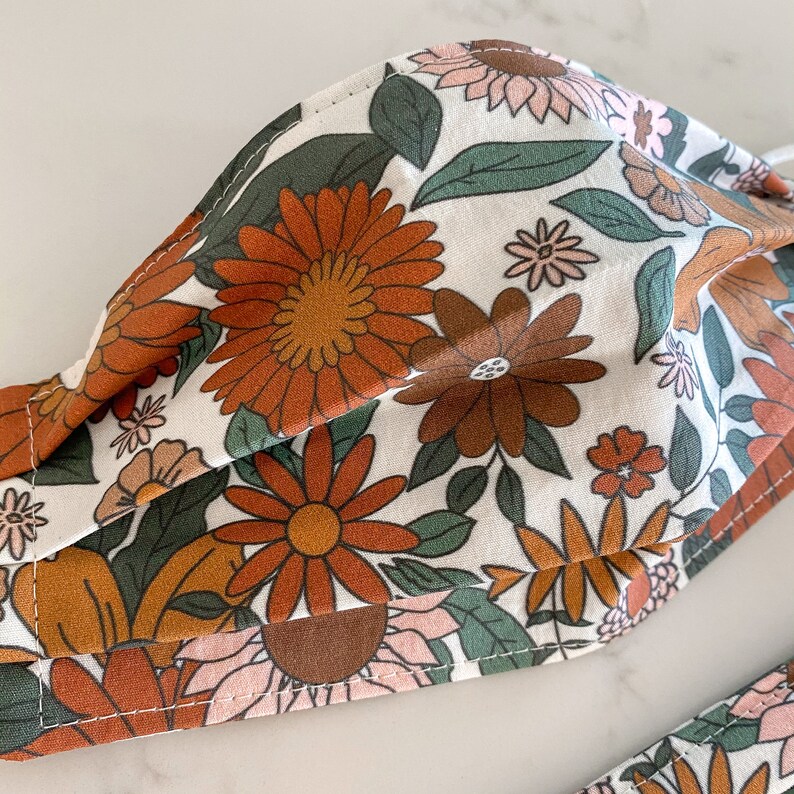 Peut inclure: Un masque facial en tissu blanc avec un motif floral compos&eacute; de fleurs orange, brunes et vertes. Le masque a un design pliss&eacute; et est cousu avec un fil brun.