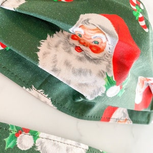 May include: Two green fabric face masks with a Christmas pattern featuring Santa Claus and candy canes. The masks are folded and have a white background with red and green accents.