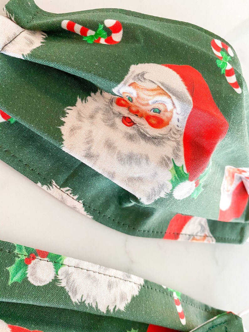May include: Two green fabric face masks with a Christmas pattern featuring Santa Claus and candy canes. The masks are folded and have a white background with red and green accents.