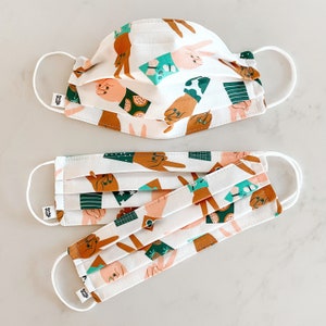 May include: Two white face masks with a repeating pattern of hands making peace signs and other abstract shapes in shades of green, brown, and blue. The masks are made of cotton fabric and have adjustable ear loops.