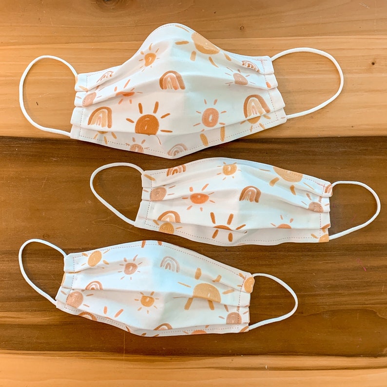 May include: Three white face masks with a pattern of suns and rainbows in shades of orange and brown. The masks have white elastic straps.