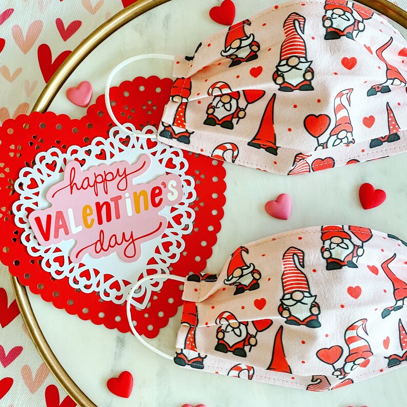 May include: Two pink fabric face masks with a pattern of gnomes and hearts. The masks have elastic ear loops. The text "happy Valentine's day" is on a red heart-shaped paper doily.