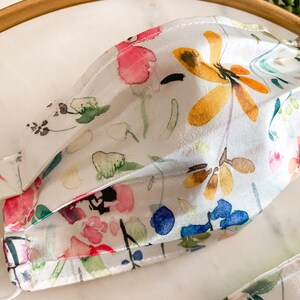 May include: A white face mask with a watercolor floral print featuring pink, yellow, blue, and green flowers.