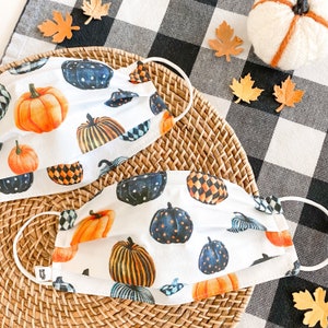 Pu&ograve; includere: Due maschere facciali bianche con un motivo a scacchi bianco e nero e un design a forma di zucca. Le maschere sono su una superficie di vimini intrecciato con uno sfondo a scacchi bianco e nero.