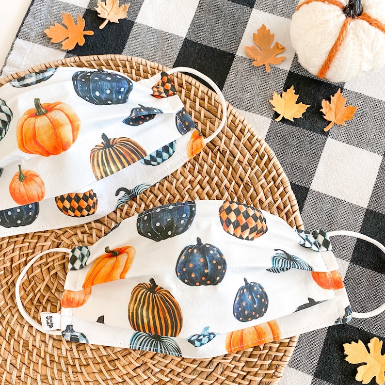 Pu&ograve; includere: Due maschere facciali bianche con un motivo a scacchi bianco e nero e un design a forma di zucca. Le maschere sono su una superficie di vimini intrecciato con uno sfondo a scacchi bianco e nero.
