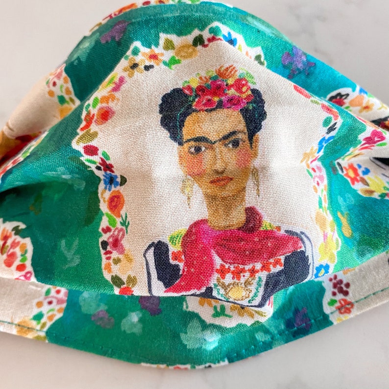 May include: A green fabric face mask with a white floral pattern and a portrait of Frida Kahlo in the center. The portrait shows Frida Kahlo with a red scarf and a flower crown.