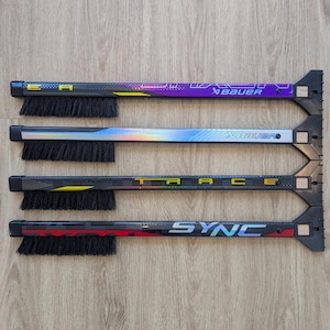 May include: Four hockey stick accessories with brushes and ice scrapers. The sticks have black, purple, silver, and red designs with the words "EA", "Bauer", "TRACE", and "SYNC". The brushes are black.