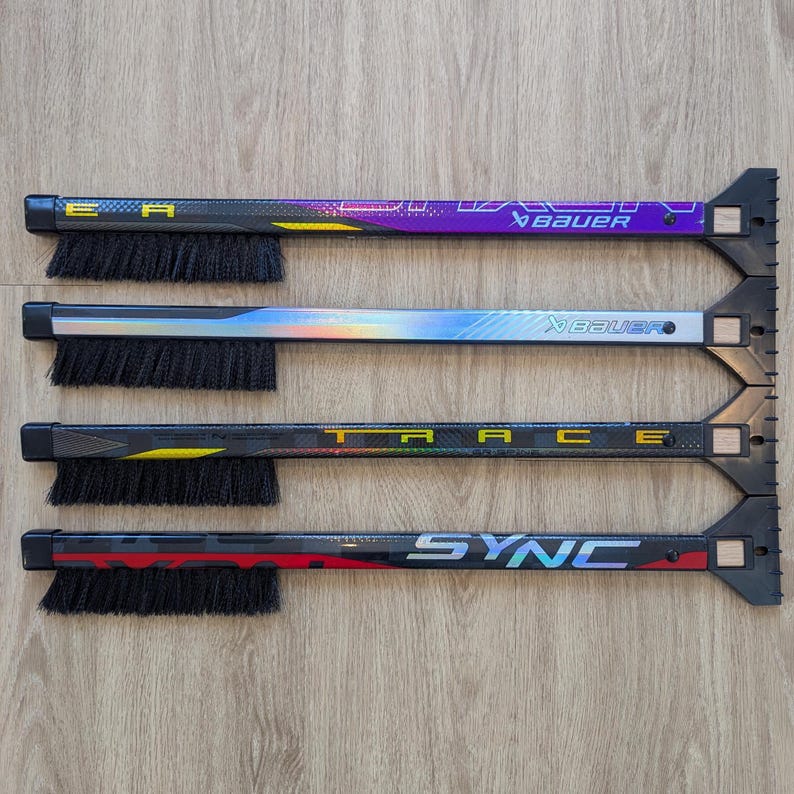 May include: Four hockey stick accessories with brushes and ice scrapers. The sticks have black, purple, silver, and red designs with the words "EA", "Bauer", "TRACE", and "SYNC". The brushes are black.