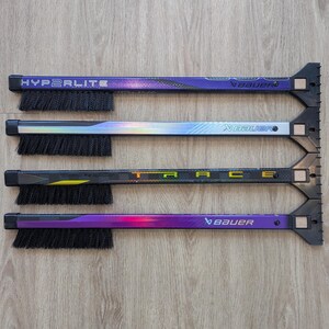 May include: Four hockey stick cleaning tools with black bristles and a scraper end. The sticks have the brand name "Bauer" and the model names "Hyperlite" and "Trace" printed on them. The sticks are purple, black, and silver.