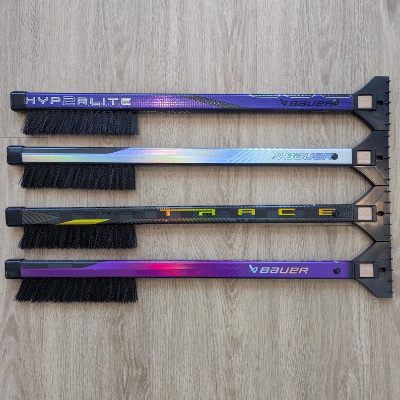 May include: Four hockey stick cleaning tools with black bristles and a scraper end. The sticks have the brand name "Bauer" and the model names "Hyperlite" and "Trace" printed on them. The sticks are purple, black, and silver.