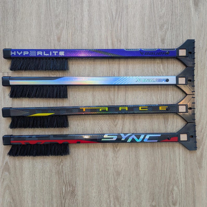May include: Four hockey stick-shaped ice scrapers with integrated brushes. The scrapers have black bristles and are labeled "HYPERLITE", "TRACE", and "SYNC". The handles are black, blue, and silver with the brand name "Bauer".